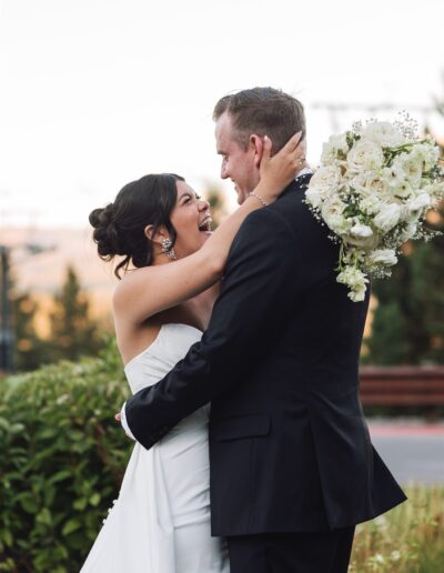 A bride and groom embrace outdoors, with the bride holding a bouquet of white flowers.