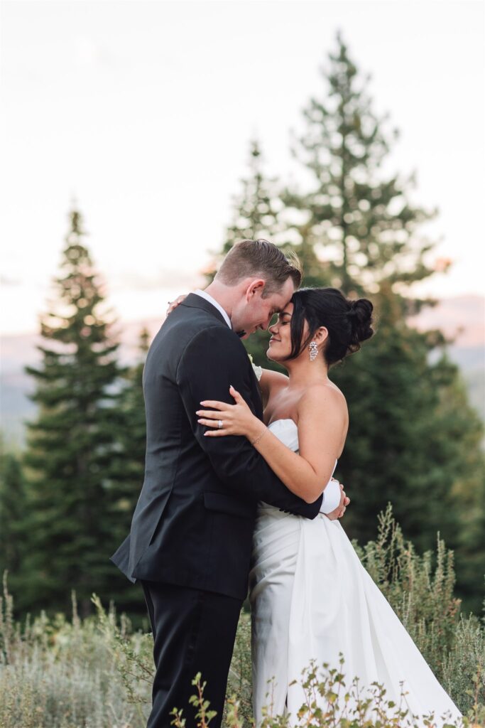 A couple wearing formal attire embraces in a forest setting with tall trees in the background.