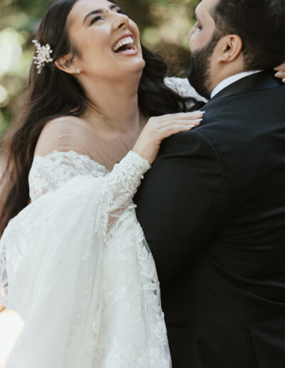 Bride and groom laughing together, outdoors. Bride in lace gown with off-shoulder design, groom in black suit. Lush greenery in the background.