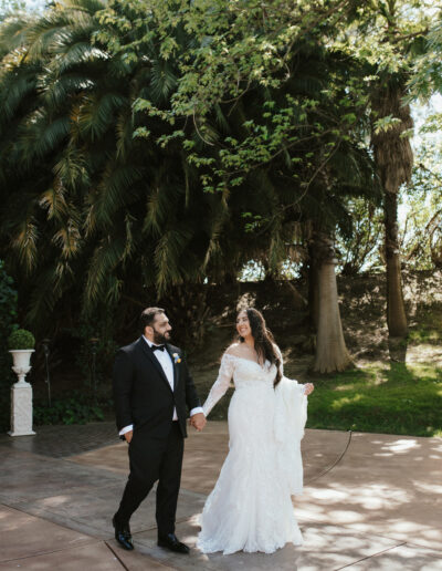 A bride and groom walk hand in hand outdoors, surrounded by lush greenery. The bride wears a white lace gown, and the groom is in a black tuxedo.