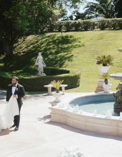 Bride and groom walk by a decorative fountain in a garden setting, surrounded by greenery and sculptures.