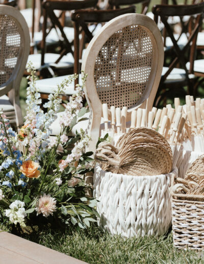 Wedding ceremony setup with wooden chairs on grass, floral arrangements, and baskets filled with woven fans and paper umbrellas.