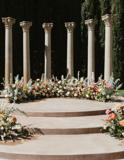 Outdoor wedding altar with a circular stone platform, decorated with colorful flower arrangements and flanked by tall columns.