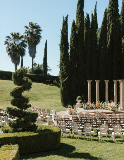 Outdoor wedding ceremony setup with chairs arranged in a semi-circle around a decorative altar, surrounded by tall trees and manicured hedges.