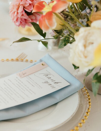 A floral centerpiece with pink and yellow flowers is next to a place setting, featuring a thank you card on a blue napkin atop a white plate with a decorative gold-rimmed edge.