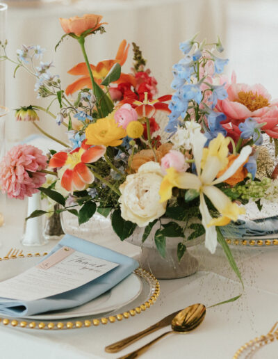 Elegant table setting with a colorful floral centerpiece, gold-rimmed glassware, and light blue napkins on white plates.