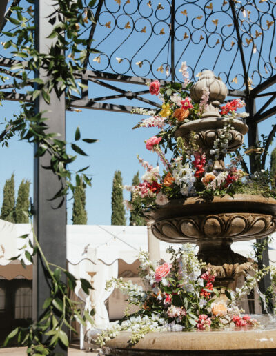 A stone fountain adorned with flowers under a metal pergola, surrounded by tents and trees, with string lights overhead.