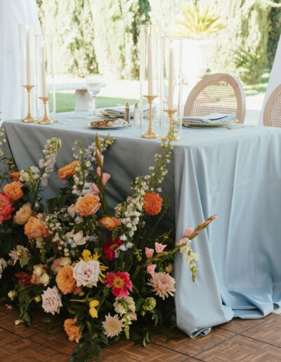 A table with a light blue tablecloth is set with candles and plates, surrounded by an arrangement of colorful flowers.