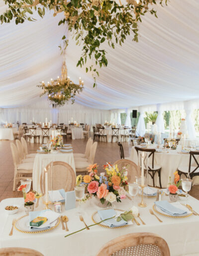 Elegant wedding reception setup under a white marquee with chandeliers, decorated tables with floral centerpieces, candles, and polished tableware.