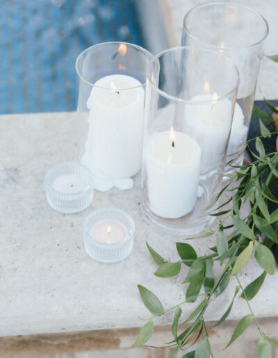 Lit white candles in glass holders on a stone ledge, with green foliage, beside a blue-tiled pool.