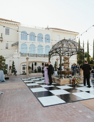 Outdoor gathering with people in formal attire around a black-and-white checkered dance floor, near a fountain, in front of a large building with arches and columns.