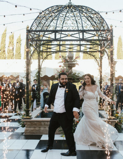 A bride and groom dance on a checkered floor under a decorative dome, surrounded by guests. Sparks rise from the floor.