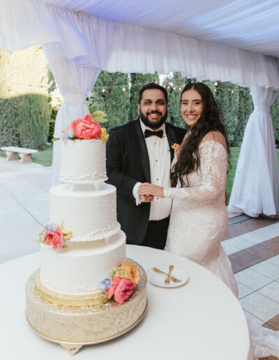 A bride and groom stand together in front of a three-tiered wedding cake with floral decorations, inside a white tent with greenery in the background.