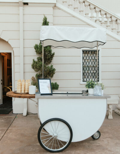 A white ice cream cart with a striped canopy is set up outdoors. Cone stacks and plant decorations are on display. A small sign is visible on the side.