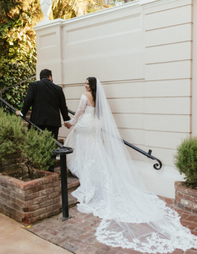 A bride in a long white dress and veil holds hands with a groom in a black suit as they ascend outdoor stairs.