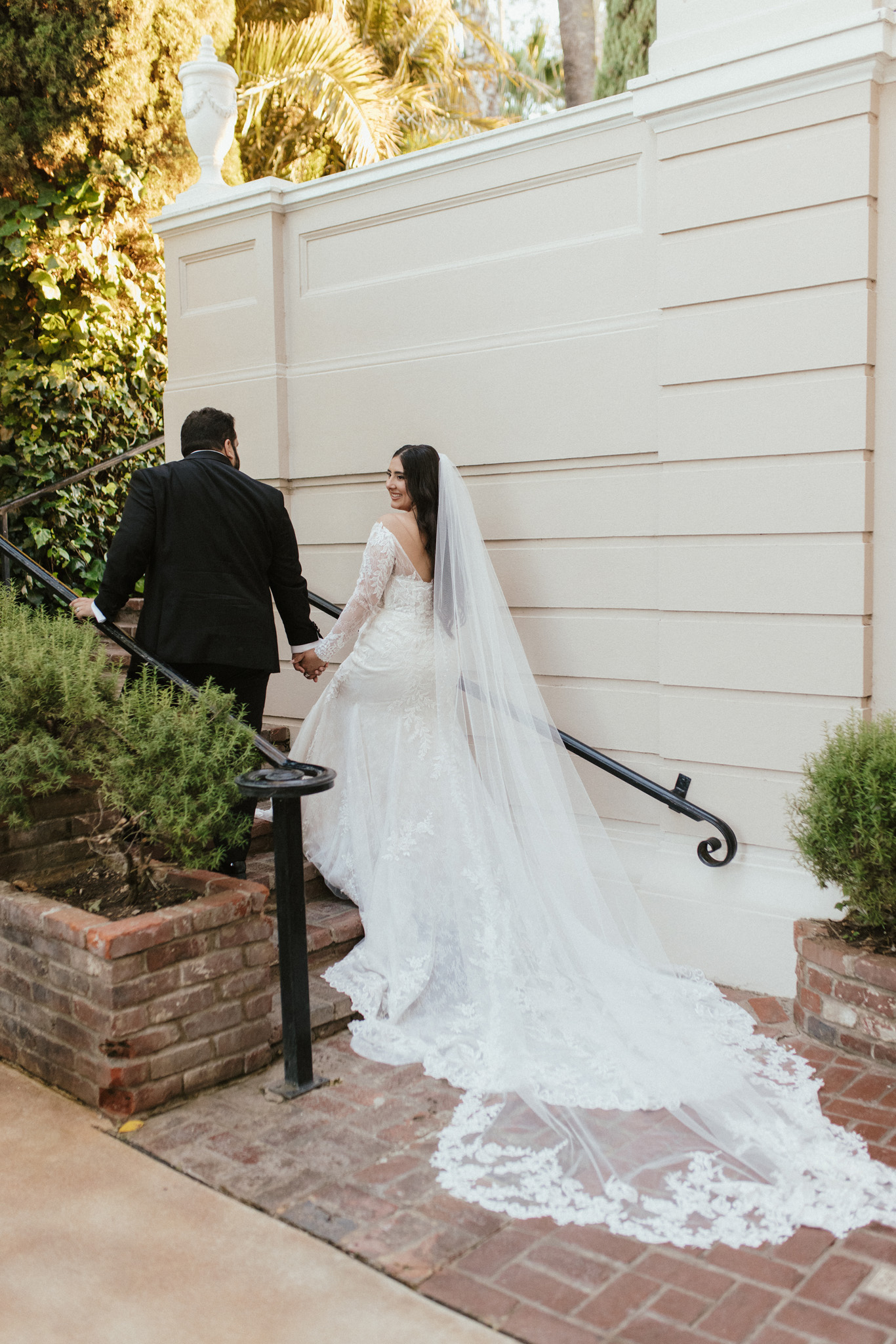 A bride in a long white dress and veil holds hands with a groom in a black suit as they ascend outdoor stairs.