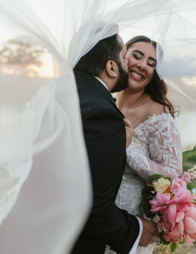 A bride and groom share a joyful moment under a flowing veil, with the bride holding pink flowers.