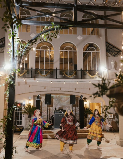 Three dancers in colorful traditional outfits perform on a checkered floor outdoors, with a grand building and decorative lights in the background.