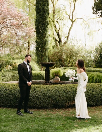 A bride and groom stand facing each other outdoors, surrounded by greenery and a hedge, with a fountain in the background. The bride holds a bouquet.