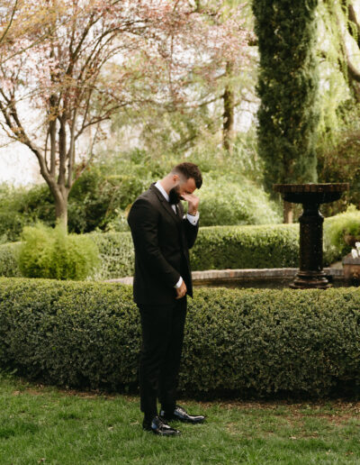 A person in a suit stands in a garden, wiping their eye. There is a fountain and trimmed hedges in the background.