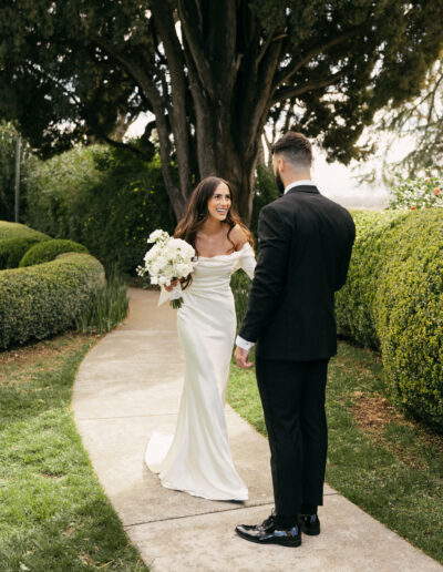 A bride in a white dress holds a bouquet, smiling at a groom in a black suit on a garden pathway. Trees and manicured bushes surround them.