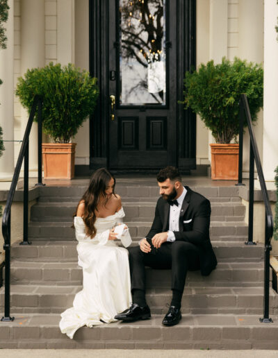 A bride and groom sit on outdoor steps in front of a black door. She wears a white gown, and he wears a black suit. Green potted plants are beside them.