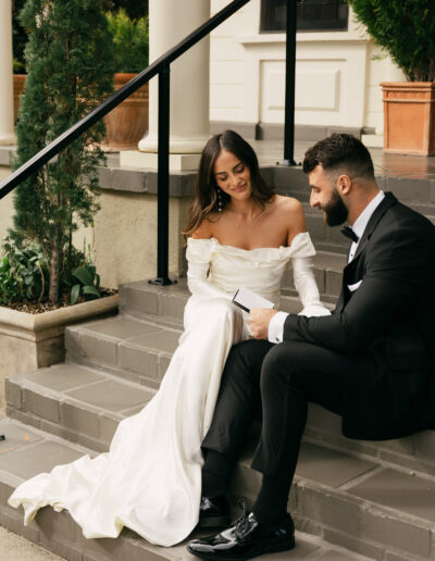 A bride in a white dress and a groom in a black tuxedo sit on steps reading from a booklet together, with plants and a building in the background.