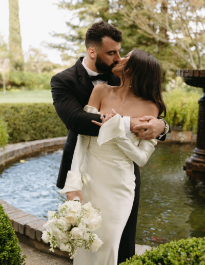 A couple in formal attire shares a kiss by a fountain in a garden. The woman holds a bouquet of white flowers.