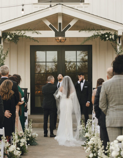 Bride and groom stand at an outdoor wedding ceremony with guests seated on either side. The bride wears a long veil. The officiant and best man are near the entrance of a building.