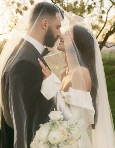 Bride and groom under a veil, smiling at each other. Bride holds a bouquet of white roses.