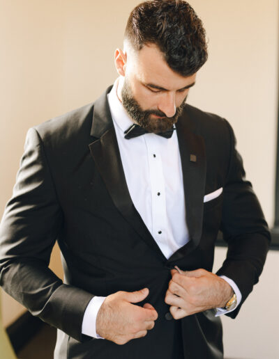 A man in a black tuxedo with a bow tie adjusts his jacket in a well-lit room.