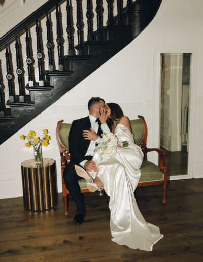 A couple in wedding attire kisses while sitting on a vintage loveseat in front of a staircase. A small table with yellow flowers is beside them.