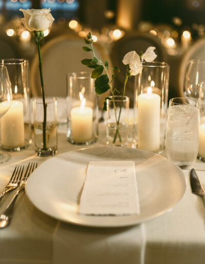 A table setting with a white plate, napkin, cutlery, and glasses. Candles in various heights and flowers in vases are arranged as centerpiece decor.