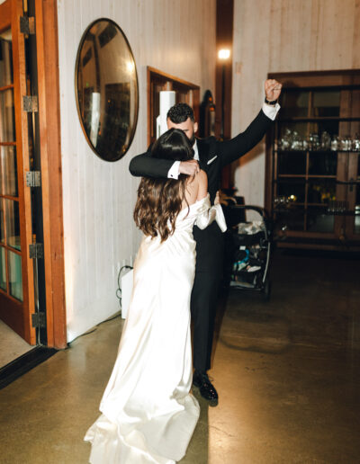 A couple dances indoors; the woman in a white dress and the man in a suit. The setting includes a round mirror and glassware on shelves.