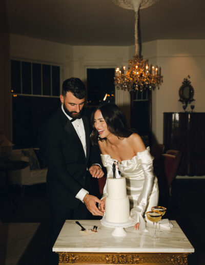 A couple in formal attire are smiling and cutting a white cake on a small table with two glasses nearby, in a dimly lit room with a chandelier.