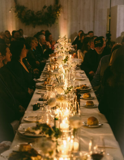 A long table set for a dinner event, with plates, glasses, and candles. People are seated on both sides, and a decorative wreath hangs on the wall in the background.