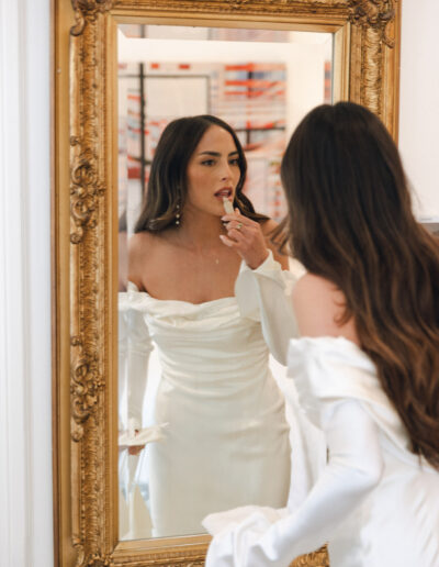 A woman in a white off-shoulder dress applies lipstick while looking in a large ornate mirror.