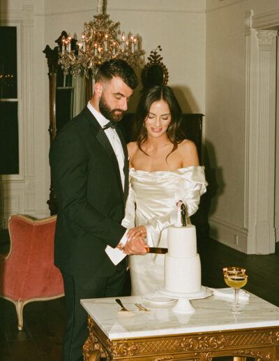 A couple in formal attire cuts a white cake on a marble table in a room with a chandelier and antique decor.