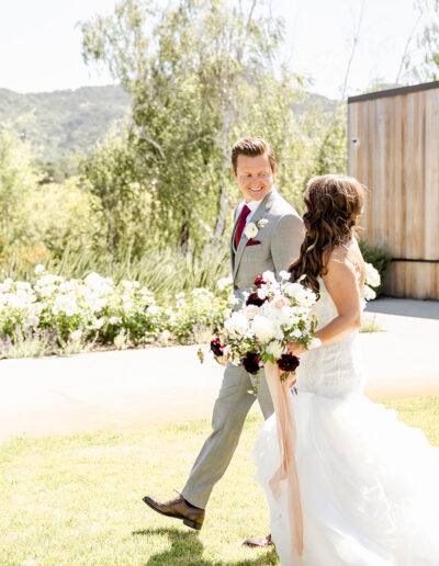 A bride and groom walk outdoors on grass near a wooden building and garden. The bride holds a bouquet of white and maroon flowers. It's a sunny day.