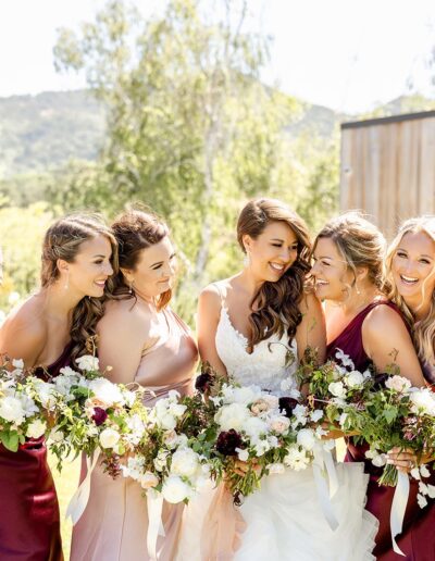 Bride surrounded by bridesmaids holding bouquets, posing outdoors in front of a scenic backdrop.