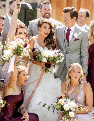 A wedding party poses joyfully; the bride and groom are surrounded by bridesmaids and groomsmen, all smiling and holding bouquets.