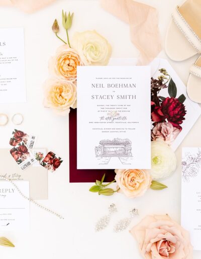 Flat lay of wedding invitation suite with floral decorations, rings, reply cards, and shoes on a white background.