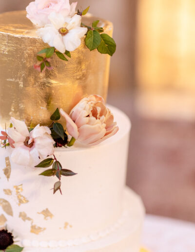 A two-tiered wedding cake with a golden top tier, adorned with white and blush flowers and green leaves.