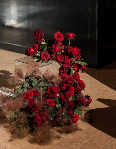 Elegant arrangement of red roses in a transparent square vase, set against a dark background with dappled shadows on a beige surface.