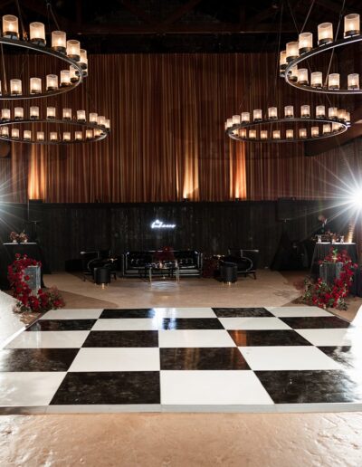 Dimly lit ballroom with black and white checkered dance floor, surrounded by brick walls, floral arrangements, and large circular chandeliers overhead.