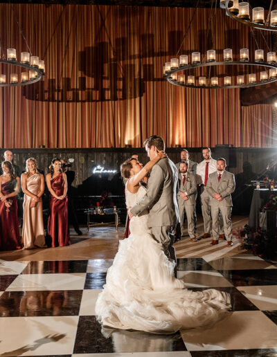 Bride and groom dance on a checkered floor at a wedding reception, surrounded by guests and lit by large circular chandeliers.