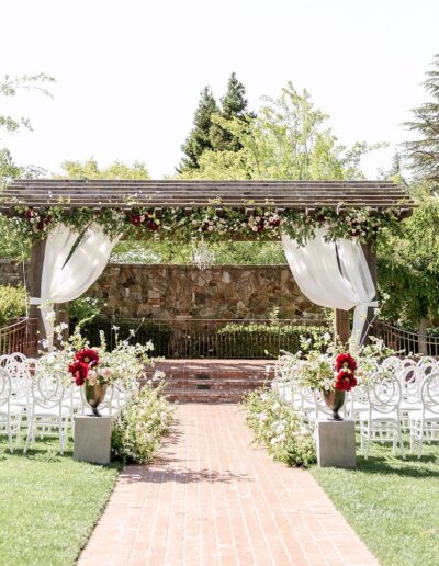 Outdoor wedding ceremony setup with white chairs, floral arrangements, and a wooden pergola draped with sheer fabric and flowers, on a grassy lawn with trees in the background.