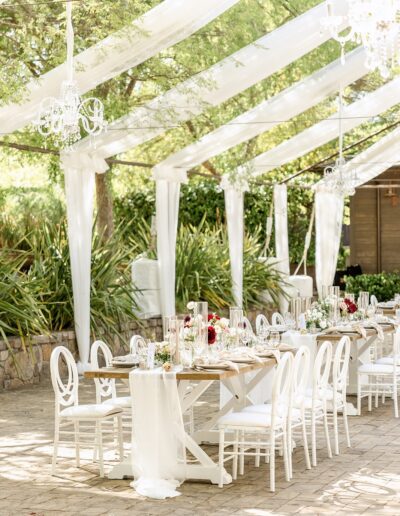 Outdoor wedding reception setup with white chairs, a long decorated table, floral centerpieces, and hanging chandeliers under a clear tent with greenery in the background.