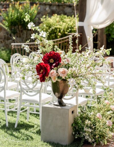 Flower arrangement with red and white blooms in a vase atop a pedestal, surrounded by greenery. Rows of white chairs are in the background, set outdoors with a draped structure.