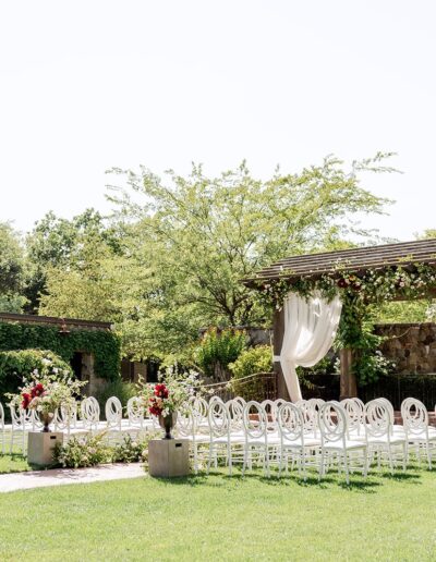 Outdoor wedding setup with white chairs arranged in rows facing a wooden pergola adorned with white drapes and flowers. Greenery surrounds the scene on a sunny day.